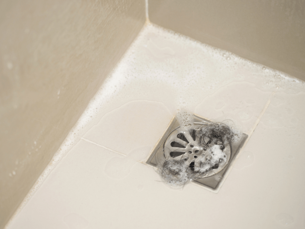 Ralo metálico de banheiro com cabelos e espuma acumulados na abertura, mostrando resíduos que podem dificultar o escoamento da água.
