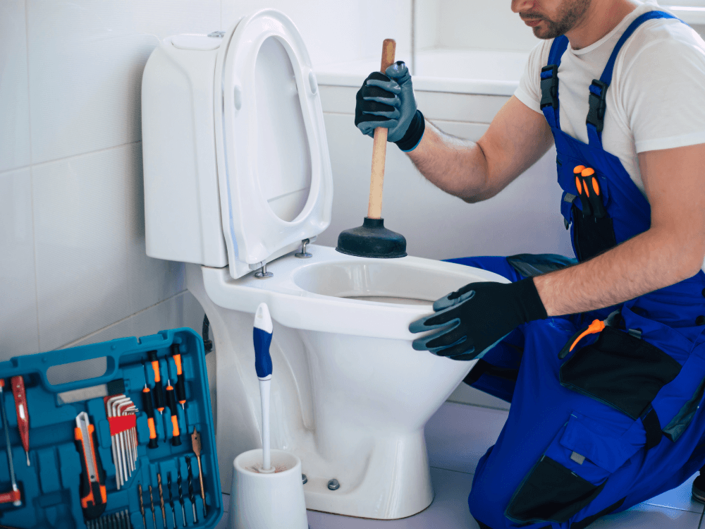Encanador utiliza um desentupidor de borracha em um vaso sanitário com a tampa levantada para solucionar o entupimento. Tudo para explicar sobre vaso entupido o que fazer.